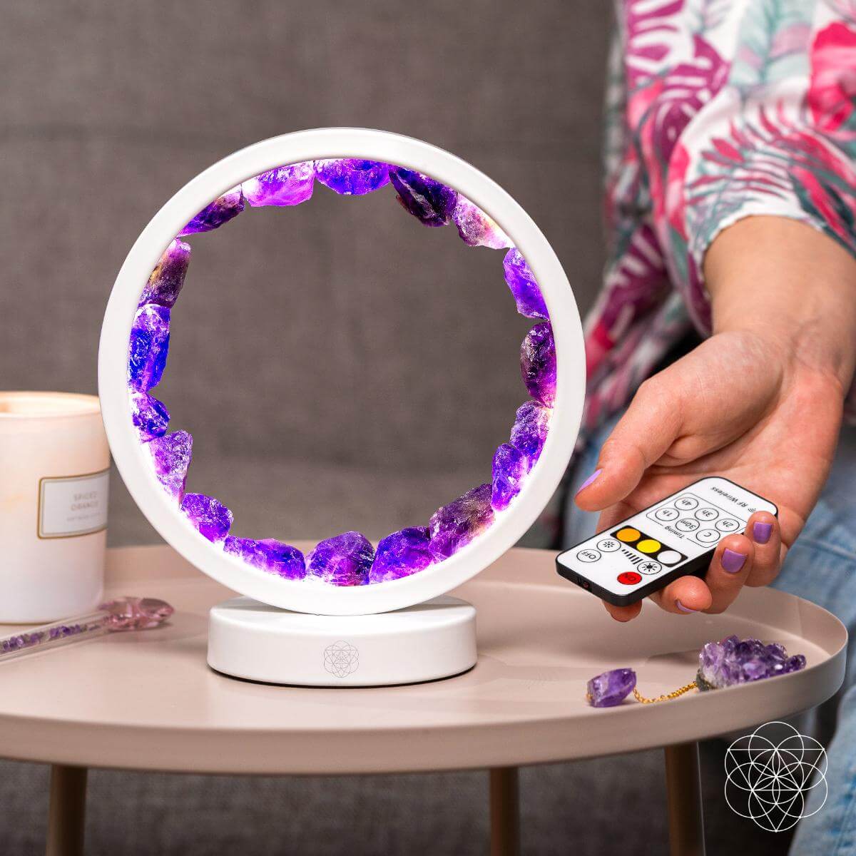The Stress Less, Sleep More – Amethyst Healing Lamp by Conscious Items glows on a table with purple crystals. A hand holds a remote by the meditation lamp, surrounded by a candle and crystals. A gray sofa sits in the background.