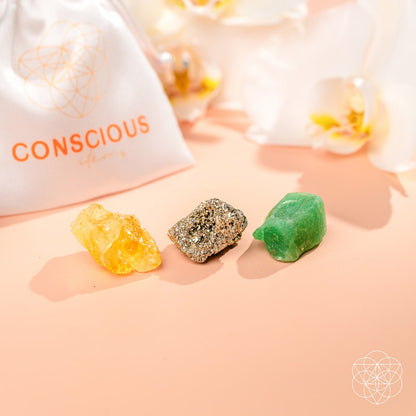 The Abundance Trio by Conscious Items features Citrine for manifestation, metallic gray pyrite for confidence, and green aventurine for abundance, displayed on a peach surface with a white drawstring bag and white flowers in the background.