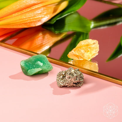 The Abundance Trio by Conscious Items features three crystals—green aventurine for abundance, yellow citrine for manifestation, and metallic pyrite for confidence—displayed on a pink surface beside a mirror with an orange flower.