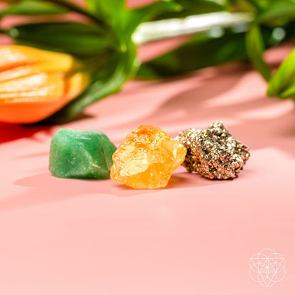 The Abundance Trio by Conscious Items—green aventurine, citrine, and pyrite crystals—is displayed on a pink surface with a blurred orange flower and green leaves in the background. A geometric white logo appears in the corner.