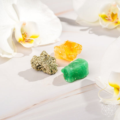The Abundance Trio by Conscious Items features golden pyrite, yellow citrine, and green aventurine crystals arranged on a light surface with white orchids. Gentle lighting adds a peaceful, elegant touch to this vibrant set.