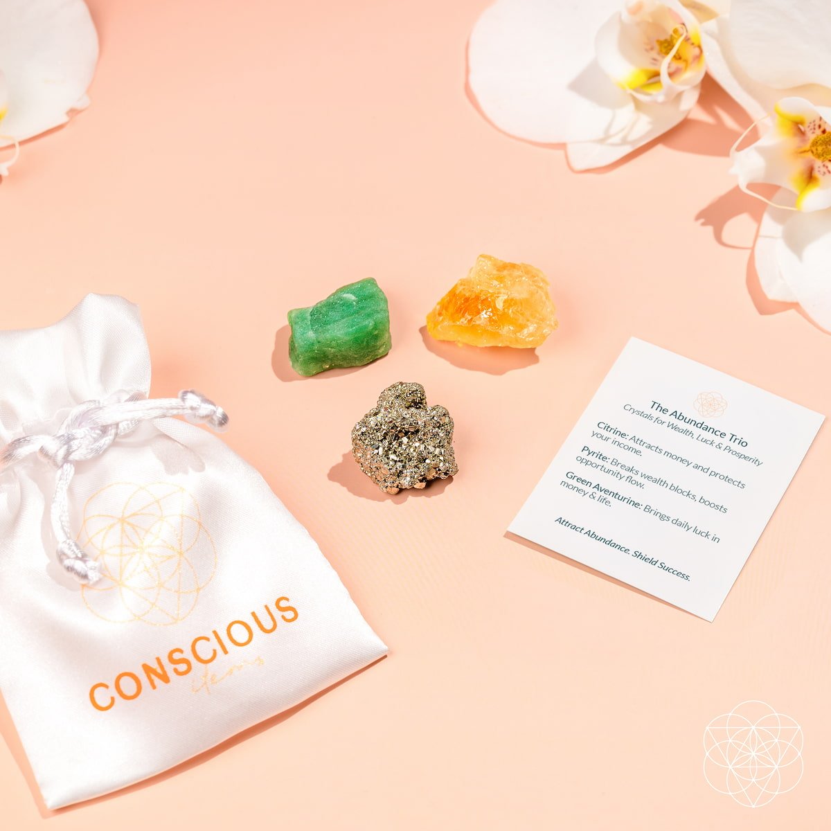 The Abundance Trio by Conscious Items includes a white silk pouch labeled CONSCIOUS on a peach surface, three crystals—green aventurine for abundance, citrine for manifestation, pyrite for confidence—an info card, and white orchids in the background.