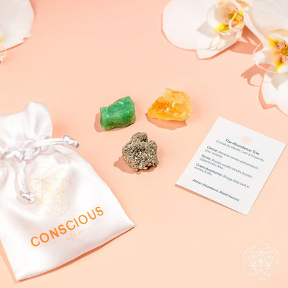 The Abundance Trio by Conscious Items includes a white silk pouch labeled CONSCIOUS on a peach surface, three crystals—green aventurine for abundance, citrine for manifestation, pyrite for confidence—an info card, and white orchids in the background.
