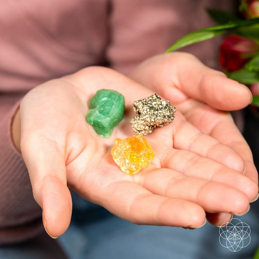 A person holds The Abundance Trio by Conscious Items—green aventurine, pyrite, and citrine crystals—in their open palm with a softly blurred background and a glimpse of a plant.
