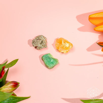 The Abundance Trio by Conscious Items—featuring Citrine, Green Aventurine, and Silver Pyrite crystals—rests on a pink surface with orange lilies and green leaves. A white geometric logo appears in the bottom right corner.
