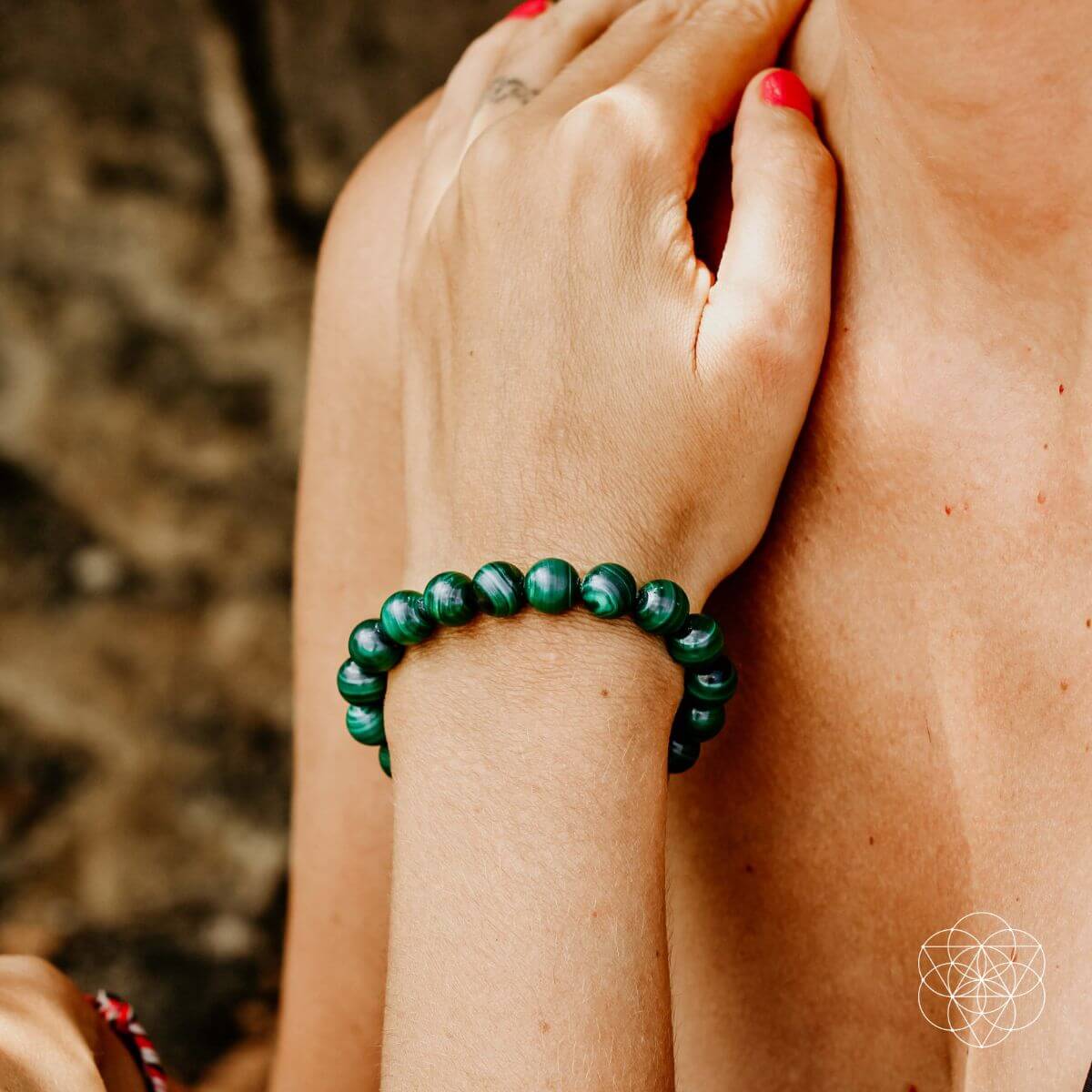 A person with pink-painted nails wears a green beaded bracelet from the Peace & Protection Bracelet Collection by Conscious Items on their wrist, hand gently resting on their upper chest against a blurred, earthy-toned background.