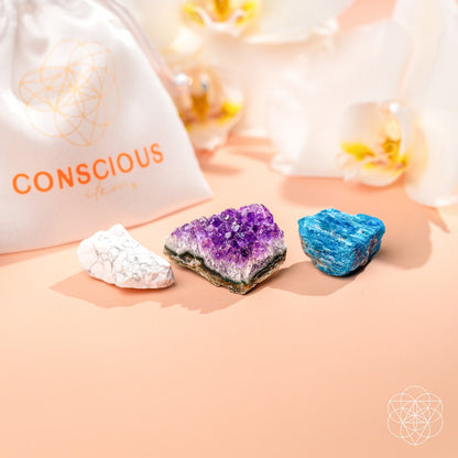 The Anti-Anxiety Trio by Conscious Items includes white, blue, and purple amethyst crystals on a peach surface next to a branded white drawstring bag, perfect for stress relief, with soft white flowers in the background.