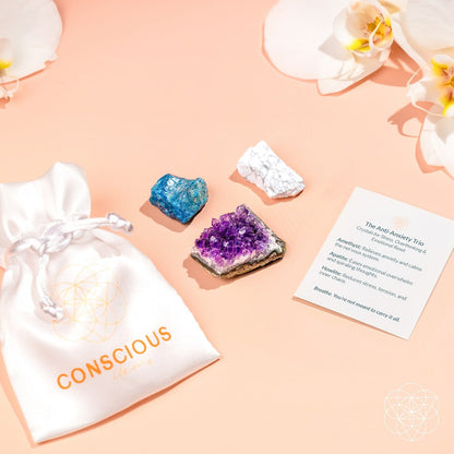 The Anti-Anxiety Trio from Conscious Items includes a silk pouch labeled “CONSCIOUS,” three calming stones (blue, white, purple), and an info card—all displayed on a peach surface with orchids, ideal for stress relief.