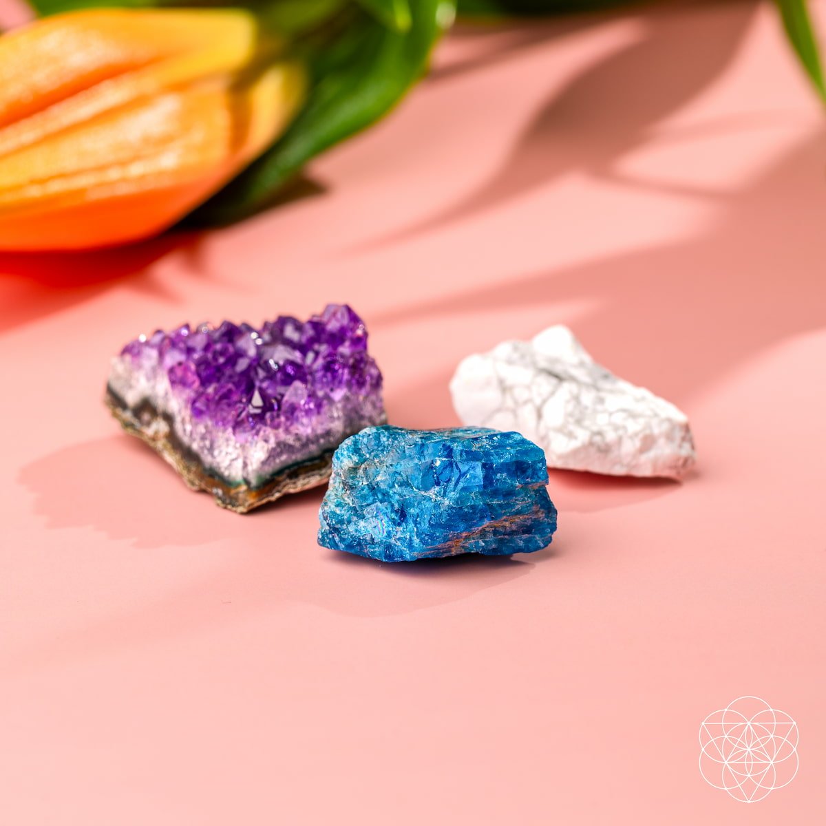 The Anti-Anxiety Trio by Conscious Items—featuring amethyst, blue mineral, and white stone—is displayed on a pink surface with an orange lily in the background. The Conscious Items geometric logo appears in the lower right corner.