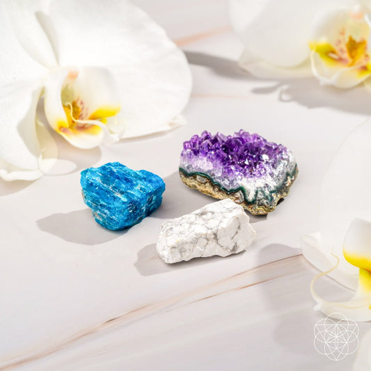 The Anti-Anxiety Trio by Conscious Items features blue, purple, and white calming stones arranged on a light surface with white orchid flowers. A soft, minimal background and geometric logo evoke stress relief and tranquility.