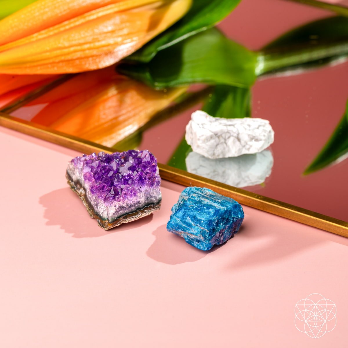 The Anti-Anxiety Trio by Conscious Items—three vibrant crystals in purple, blue, and white—rests on a pink surface near a mirror reflecting an orange lily and green leaves. A geometric white Conscious Items logo appears at the lower right.