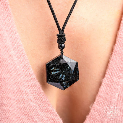 A close-up of a person wearing The Arfvedsonite Deep Healing Pendant by Conscious Items on a black cord necklace. The hexagonal black crystal rests against their chest, complementing their pink V-neck top.