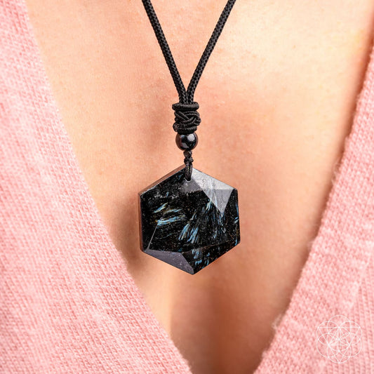 A close-up of a person wearing The Arfvedsonite Deep Healing Pendant by Conscious Items on a black cord necklace. The hexagonal black crystal rests against their chest, complementing their pink V-neck top.