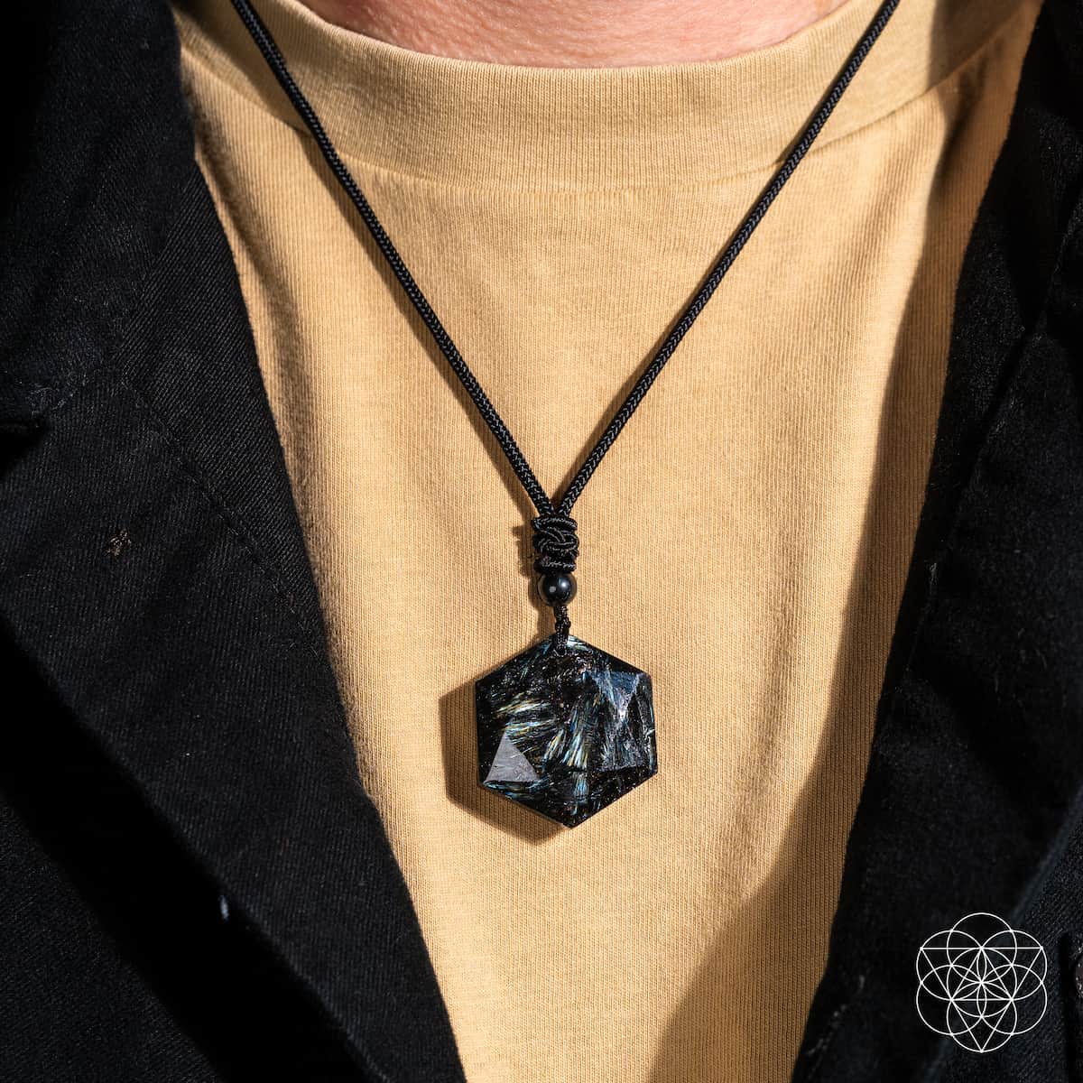 A close-up shows a person wearing Conscious Items' Arfvedsonite Deep Healing Pendant—a shimmering black cord necklace featuring the vision stone, believed to support emotional healing, styled over a tan t-shirt and black jacket.
