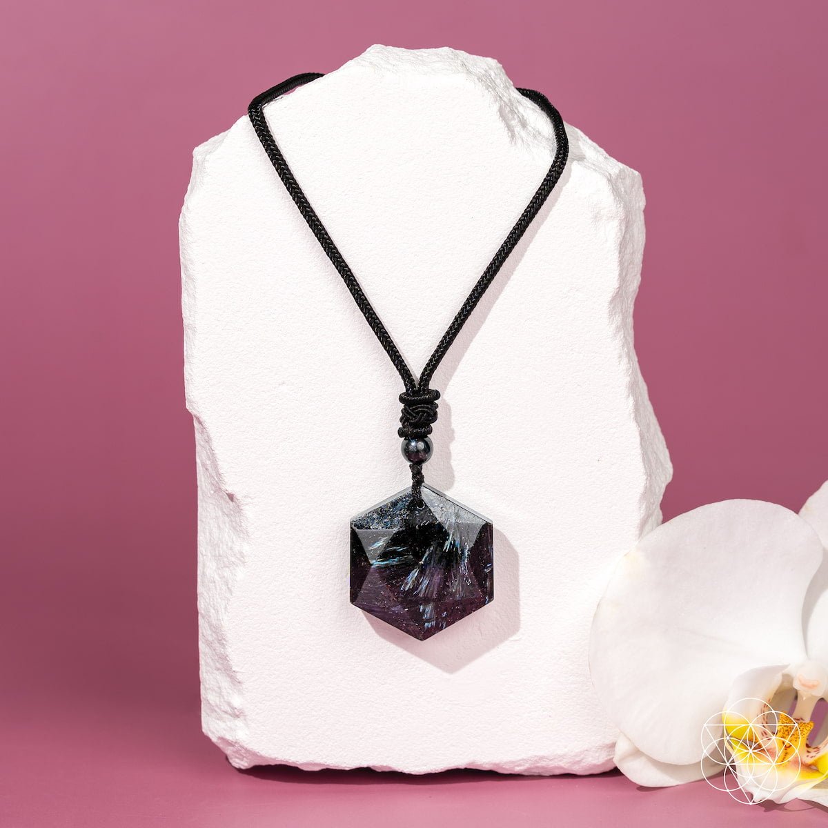The Arfvedsonite Deep Healing Pendant by Conscious Items, featuring a black crystal on a black cord, rests on a white pedestal with an orchid—an elegant vision stone believed to aid emotional healing.