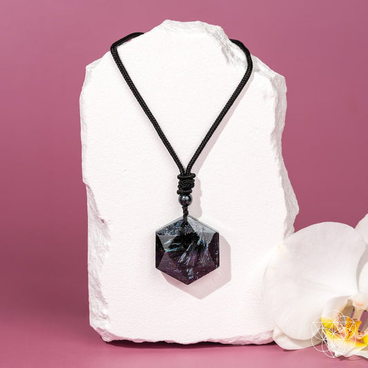 The Arfvedsonite Deep Healing Pendant by Conscious Items, featuring a black crystal on a black cord, rests on a white pedestal with an orchid—an elegant vision stone believed to aid emotional healing.