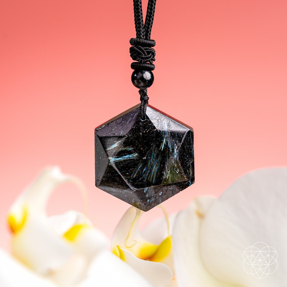 The Arfvedsonite Deep Healing Pendant by Conscious Items features a hexagonal black stone with blue-green streaks on a black cord, displayed with orchid petals—an ideal vision stone and crystal for emotional healing.