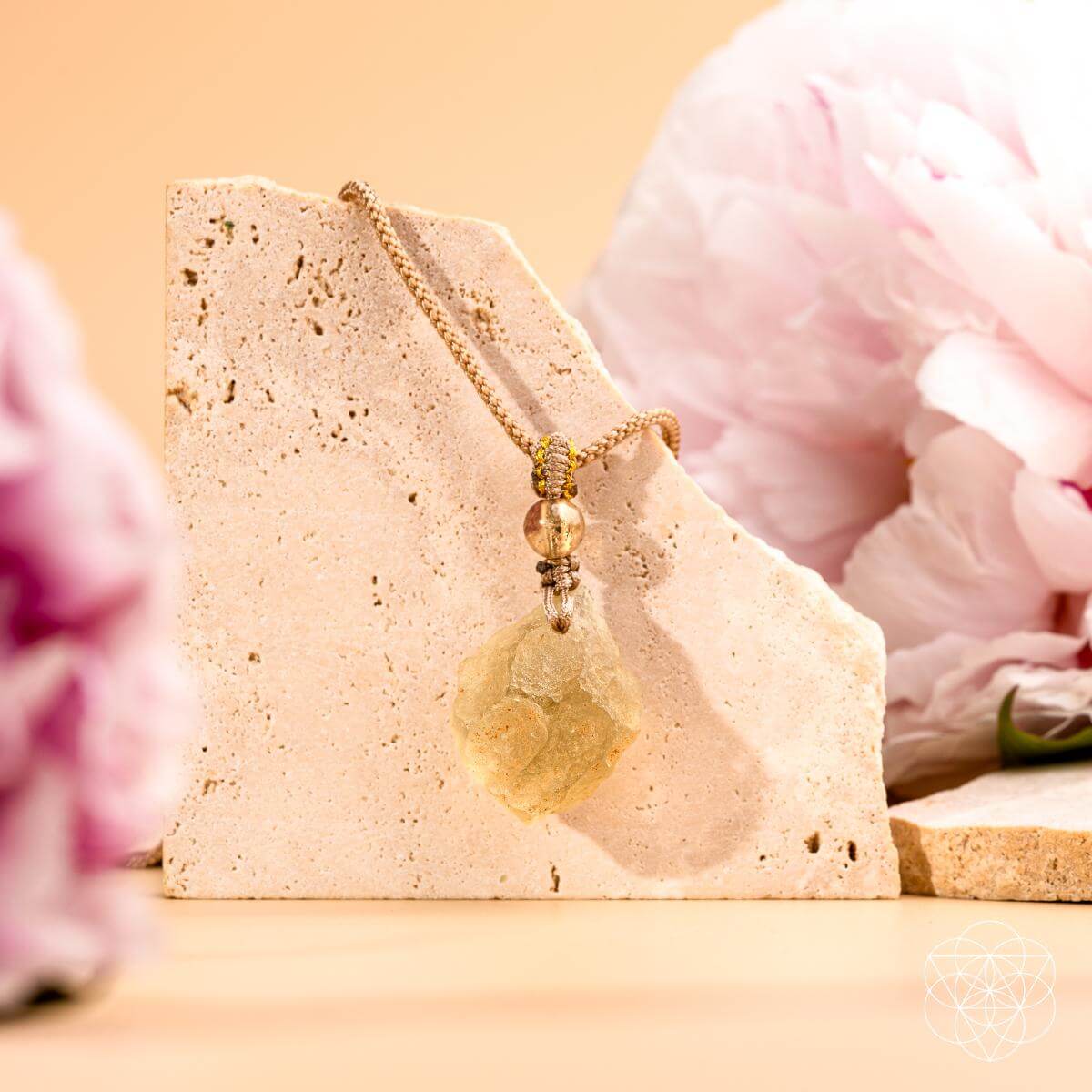 The Destiny Relic – Libyan Desert Glass Pendant by Conscious Items, set on a gold chain and shown atop a textured stone with blurred soft pink flowers, glows radiantly as a unique manifestation piece.