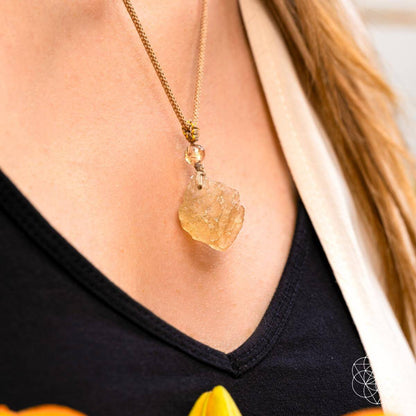 A close-up of a person wearing Conscious Items' The Destiny Relic – Libyan Desert Glass Pendant, a bold gold chain necklace, paired with a black top and cream outer garment.