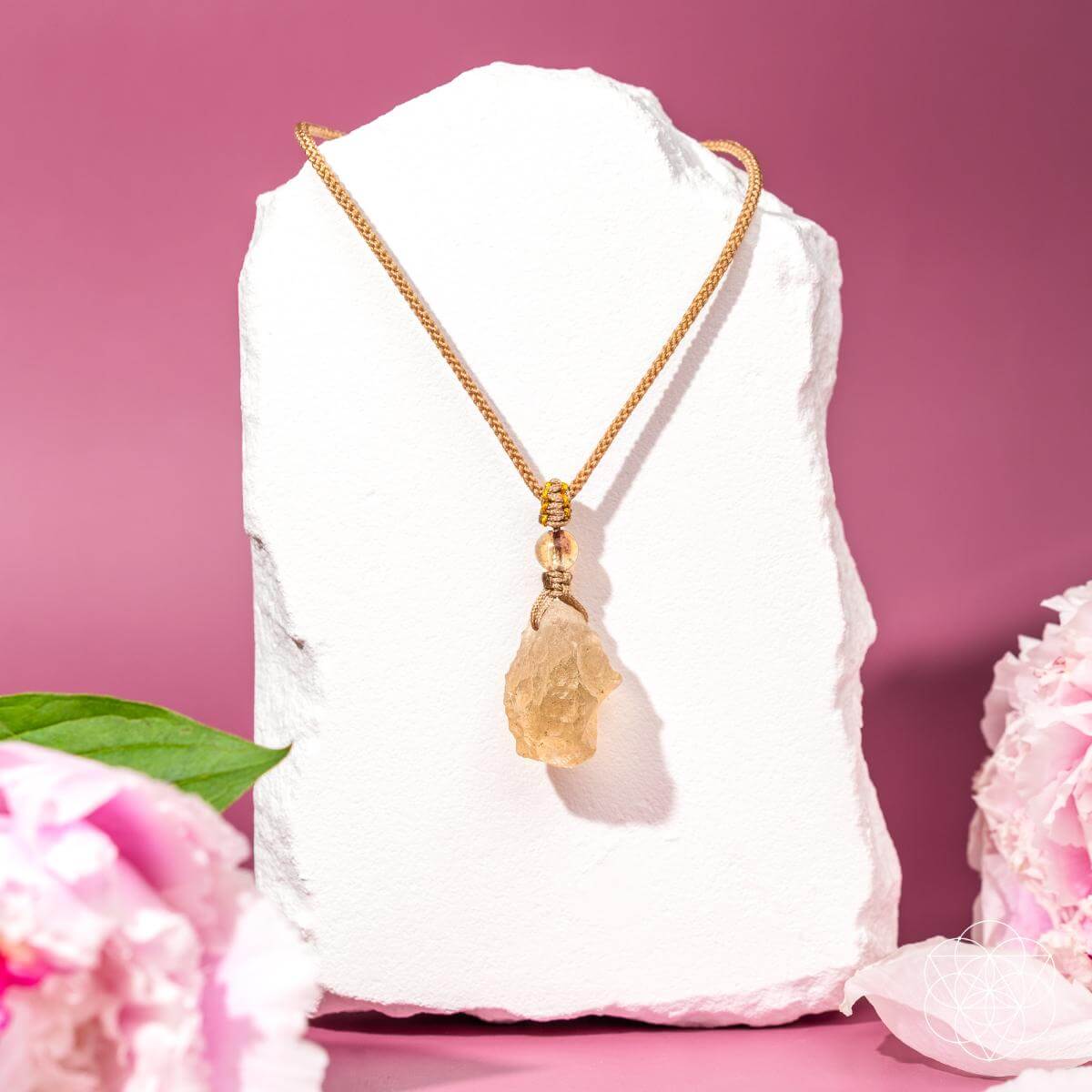 The Destiny Relic – Libyan Desert Glass Pendant by Conscious Items, a gold chain necklace featuring a rough Libyan Desert Glass pendant, is displayed on a white stone stand with a pink background, pink flowers, and green leaves.