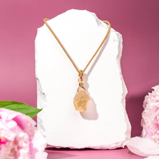 The Destiny Relic – Libyan Desert Glass Pendant by Conscious Items, a gold chain necklace featuring a rough Libyan Desert Glass pendant, is displayed on a white stone stand with a pink background, pink flowers, and green leaves.
