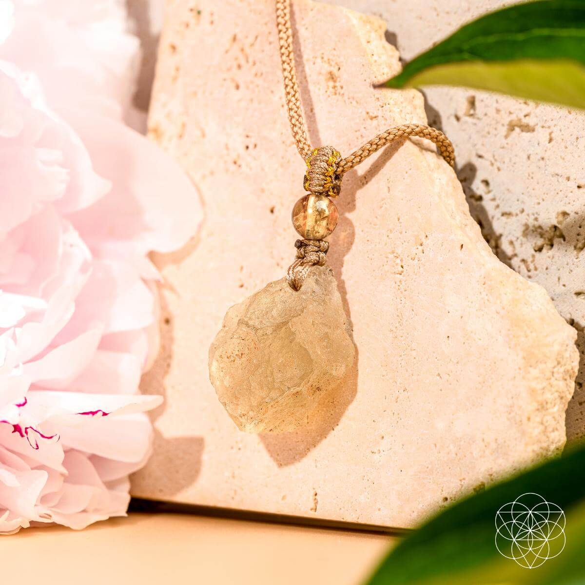 The Destiny Relic – Libyan Desert Glass Pendant by Conscious Items, featuring a raw crystal on a beige cord, rests on light stone tiles amid green leaves and pale pink flowers as warm sunlight creates soft shadows.