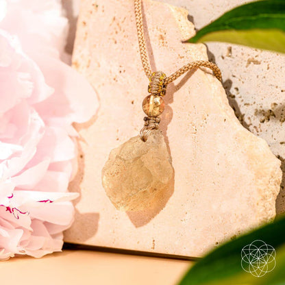 The Destiny Relic – Libyan Desert Glass Pendant by Conscious Items, featuring a raw crystal on a beige cord, rests on light stone tiles amid green leaves and pale pink flowers as warm sunlight creates soft shadows.