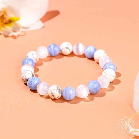 The Empath’s Trio - Daily Reset Bracelet by Conscious Items features alternating blue, white marbled, and light pink beads, arranged on a peach surface with white flower petals—ideal empath jewelry for emotional healing.