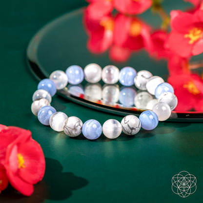 The Empath’s Trio - Daily Reset Bracelet by Conscious Items, featuring light blue, white, and marbled Blue Lace Agate stones, rests on a green surface near a mirror and red flowers. A geometric logo marks this empath jewelry in the bottom right corner.