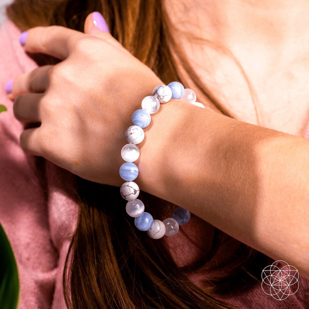 A close-up of a person with light skin, long brown hair, pink top, and lavender nails wearing The Empath’s Trio - Daily Reset Bracelet by Conscious Items, featuring white and light blue stones for emotional healing.