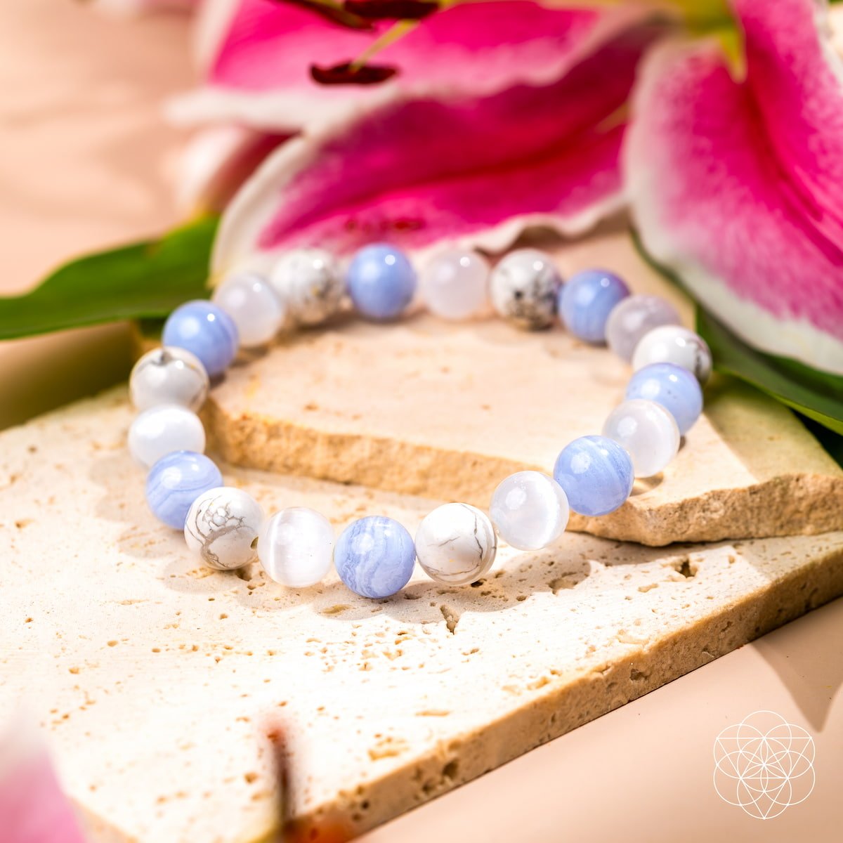 The Empath’s Trio - Daily Reset Bracelet by Conscious Items, featuring blue, white, and marbled emotional healing stones, rests on light stone tiles amid pink and white lilies in gentle natural light.