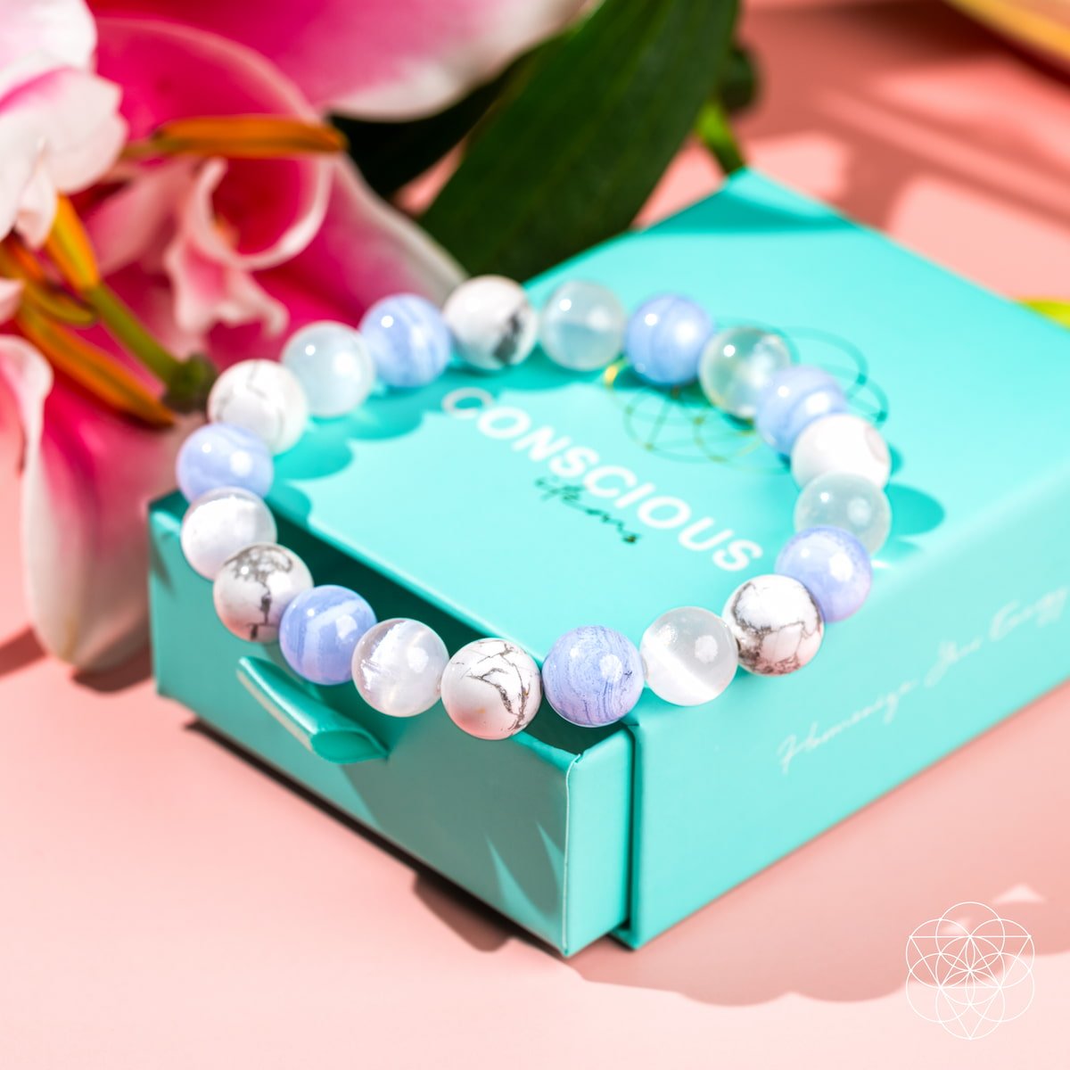 The Empath’s Trio - Daily Reset Bracelet by Conscious Items features blue, white, and marbled stones, displayed on a turquoise Conscious box with pink lilies on a light pink surface—ideal empath jewelry for emotional healing.