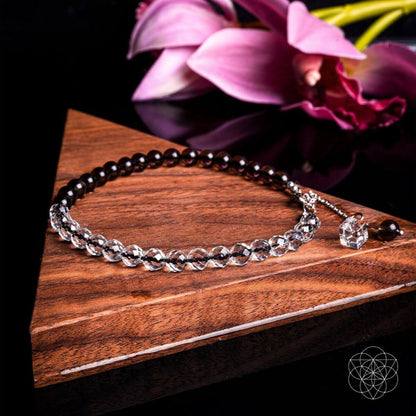 The Energy Shield Anklet by Conscious Items features alternating dark and clear faceted beads for chakra balance. Displayed on a wooden triangle, it’s set against pink lilies with a geometric logo in the corner.