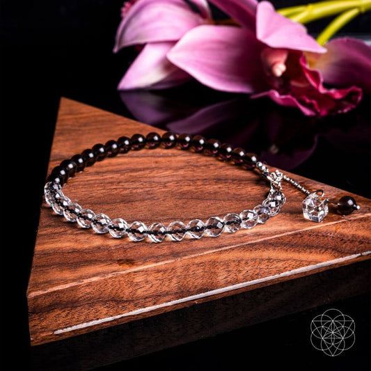 The Energy Shield Anklet by Conscious Items features alternating dark and clear faceted beads for chakra balance. Displayed on a wooden triangle, it’s set against pink lilies with a geometric logo in the corner.