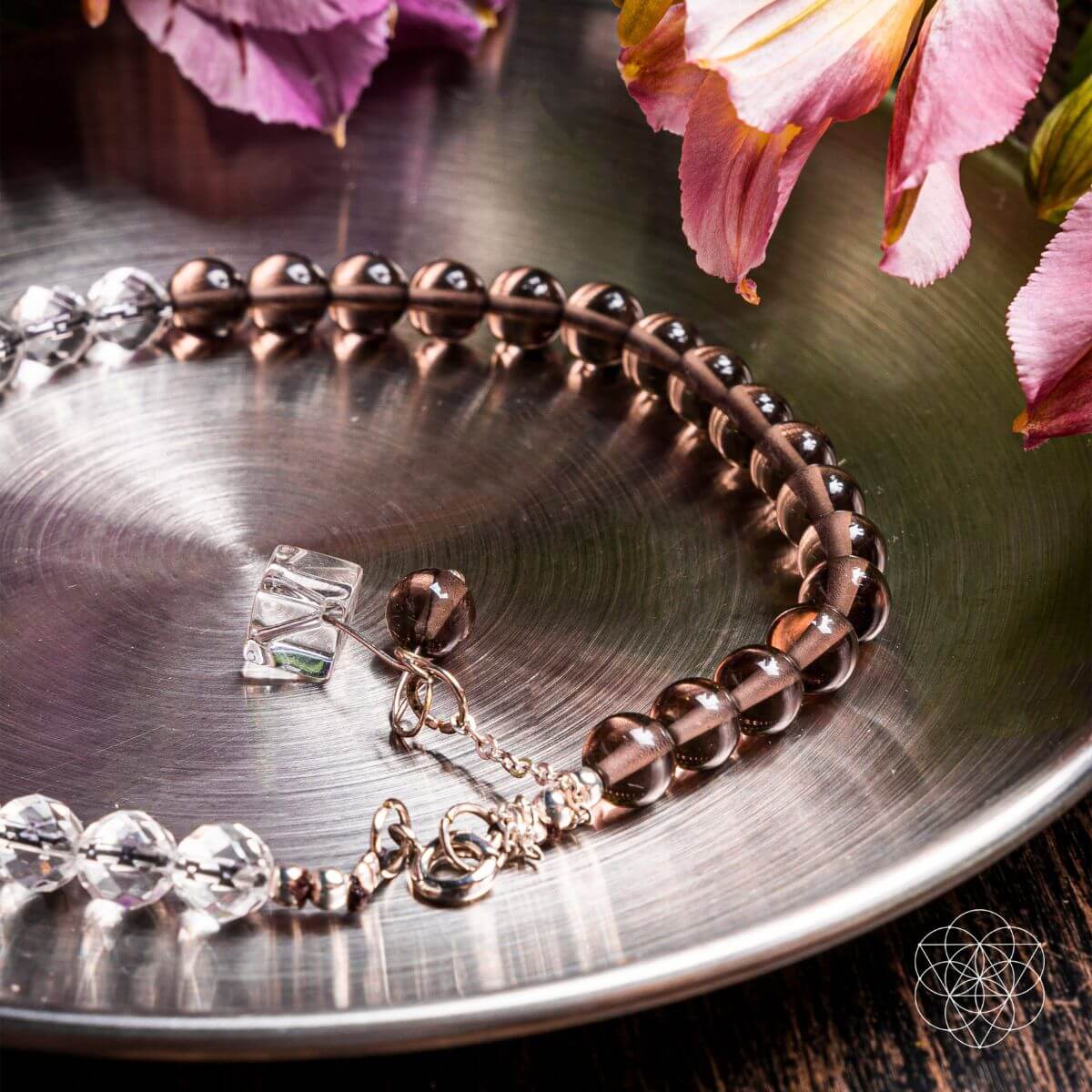 The Energy Shield Anklet by Conscious Items, featuring smoky quartz and clear crystal beads, rests on a shiny metal tray with pink flowers, evoking crystal protection. A geometric logo appears in the lower right corner.