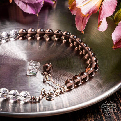 The Energy Shield Anklet by Conscious Items, featuring smoky quartz and clear crystal beads, rests on a shiny metal tray with pink flowers, evoking crystal protection. A geometric logo appears in the lower right corner.