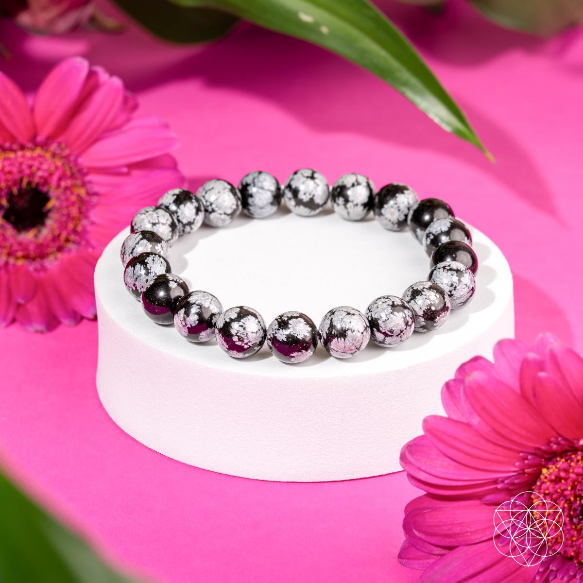 The Pattern Breaker – Snowflake Obsidian Bracelet by Conscious Items is showcased on a white stand amid bright pink flowers and green leaves against a vibrant pink background—ideal for breaking negative patterns.