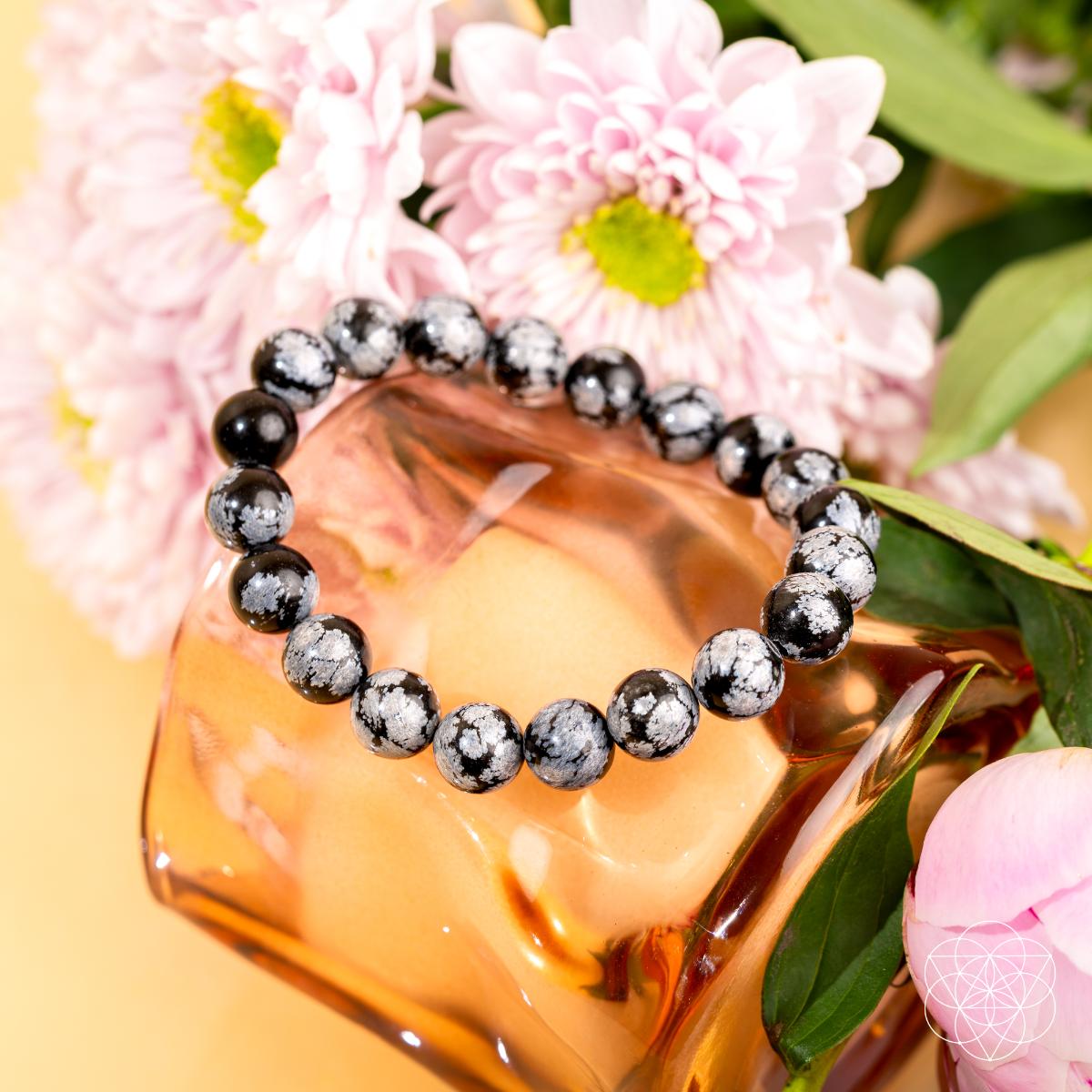 The Pattern Breaker – Snowflake Obsidian Bracelet by Conscious Items rests on a translucent pink glass vase amid pink flowers and green leaves, its presence on a light background emphasizing its power to break negative patterns.
