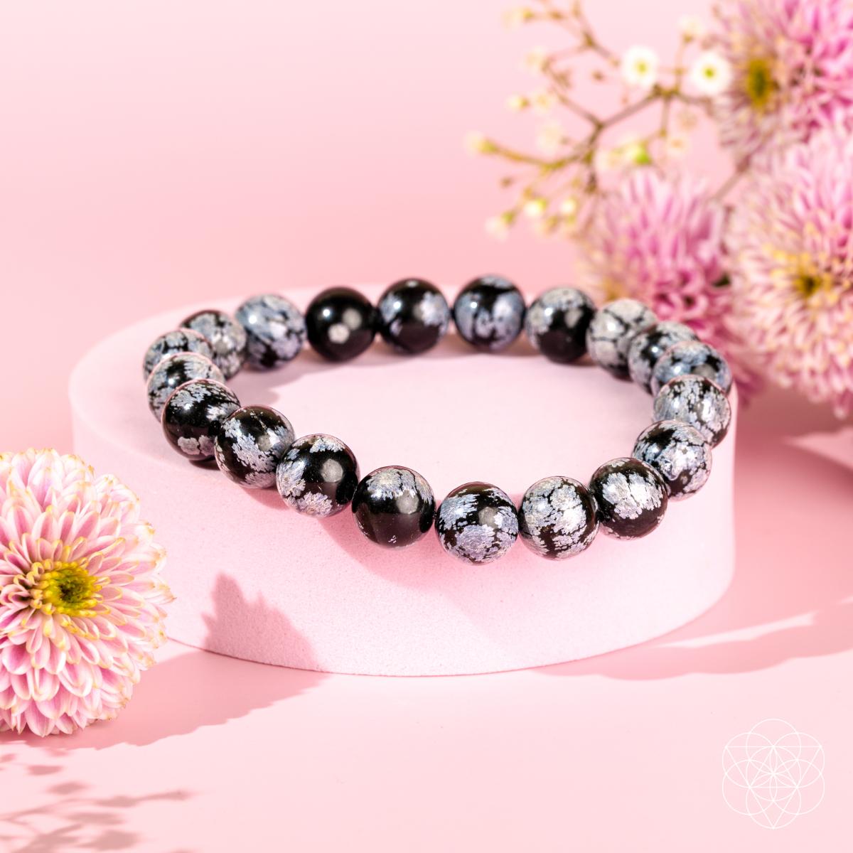 The Pattern Breaker – Snowflake Obsidian Bracelet by Conscious Items rests on a pink pedestal with floral accents, set against a soft pink background—ideal for anyone looking to break negative patterns with healing crystals.