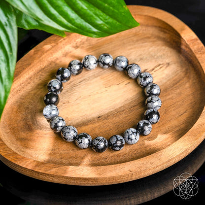 The Pattern Breaker – Snowflake Obsidian Bracelet by Conscious Items rests on a wooden tray with a green leaf nearby and a geometric logo in the corner, designed to help you break negative patterns.