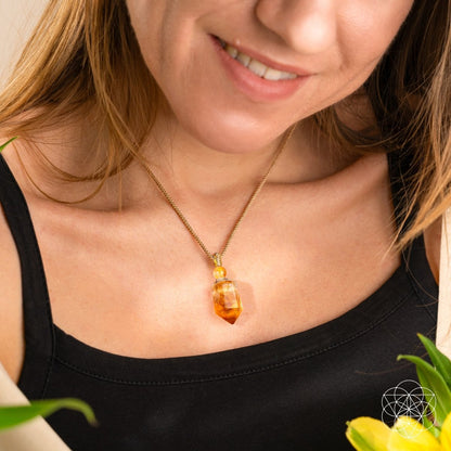 A woman in a black top and gold chain necklace featuring The Prosperity Pendant by Conscious Items—crafted with citrine, the crystal of prosperity—smiles gently. Yellow flowers and green leaves appear in the foreground.