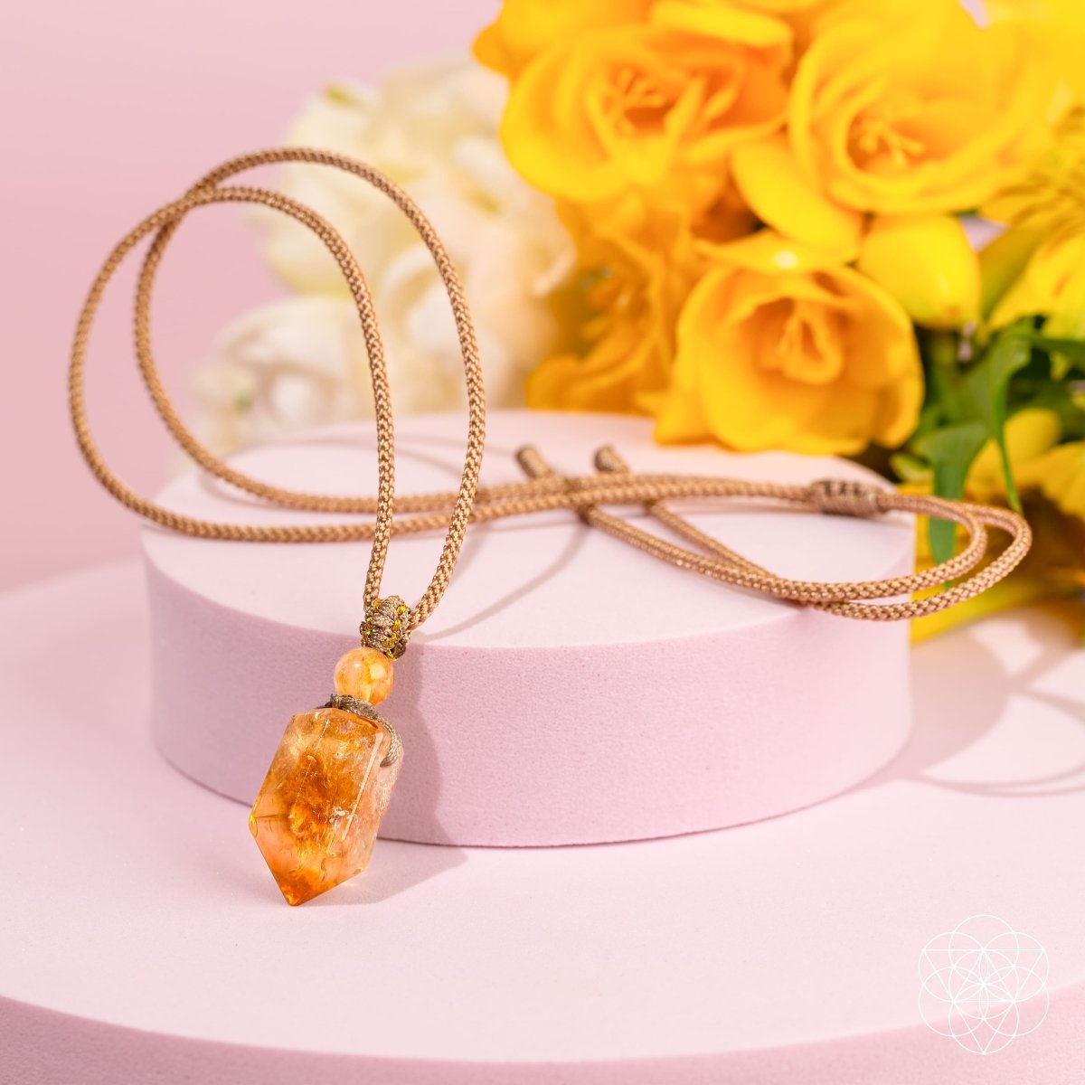 The Prosperity Pendant by Conscious Items, a crystal of prosperity on a gold chain, is displayed on a pastel pink platform with vibrant yellow flowers and a soft pink background that highlight its elegance.