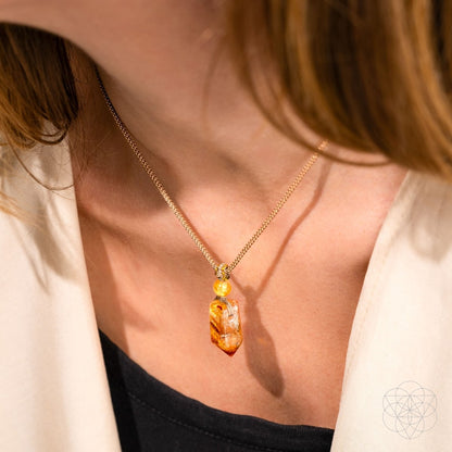 A person wears The Prosperity Pendant from Conscious Items—a gold chain necklace featuring the crystal of prosperity—styled with a light blazer and black top. The pendant and elegant lower neckline are the focus.