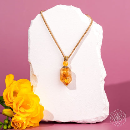 The Prosperity Pendant by Conscious Items, crafted in gold with citrine—the crystal of prosperity—rests on a white stone slab. Bright yellow flowers are to the left, set against a pink background with a geometric white logo at the bottom right.