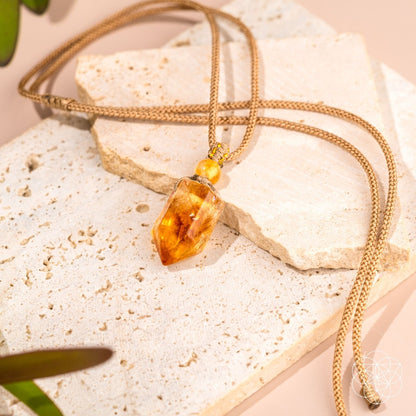The Prosperity Pendant by Conscious Items, a gold chain necklace with a faceted crystal pendant symbolizing abundance, rests on textured stone slabs against a soft beige background, accented by green leaves at the edges.