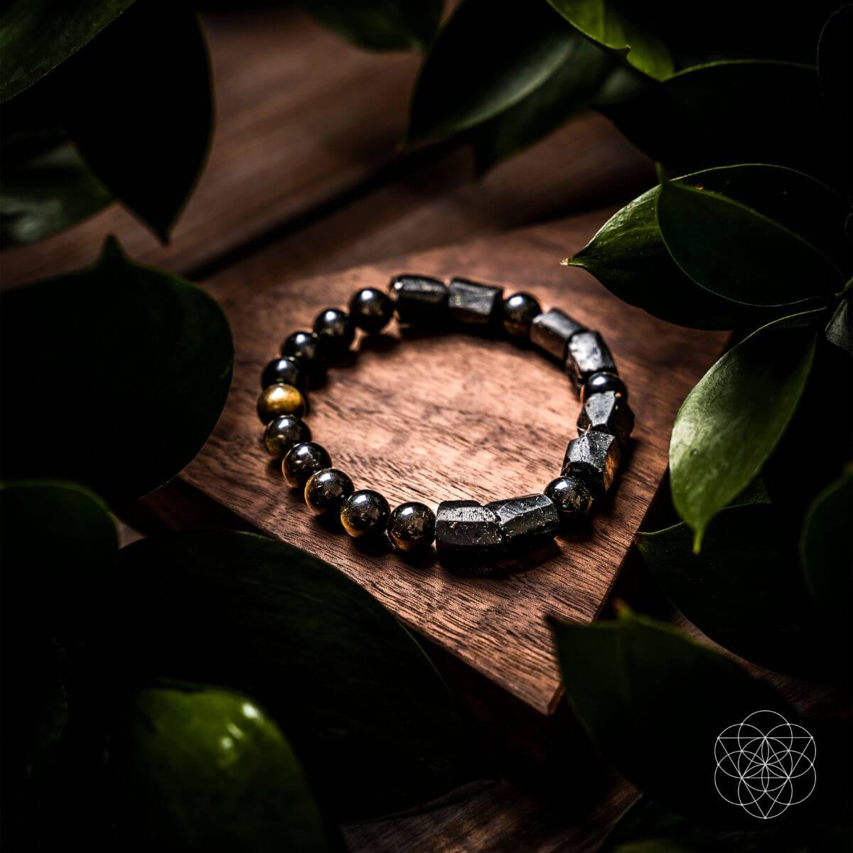 The Peace & Protection Bracelet Collection by Conscious Items, featuring dark metallic and stone-like beads, is displayed on a wooden surface amid green leaves, creating a natural and elegant look.