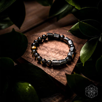 The Peace & Protection Bracelet Collection by Conscious Items, featuring dark metallic and stone-like beads, is displayed on a wooden surface amid green leaves, creating a natural and elegant look.