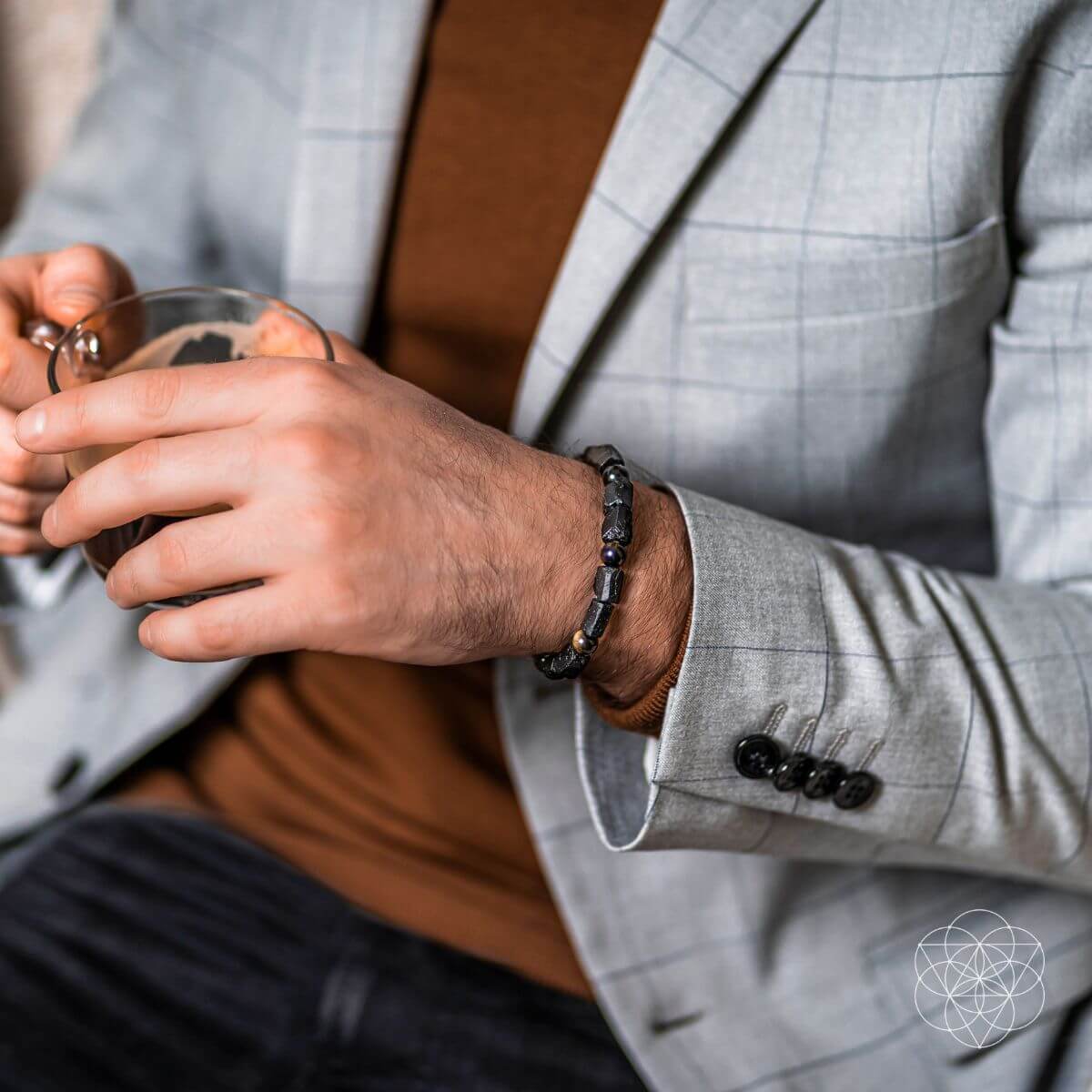 A person in a light gray plaid blazer and brown sweater holds a glass cup, wearing a Conscious Items Peace & Protection Bracelet Collection on their wrist. The image highlights their hands and torso while keeping the face out of view.