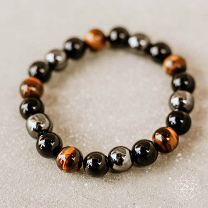 The Triple Protection Bracelet by Conscious Items showcases alternating black, silver, and brown tiger’s eye beads arranged in a circle—an elegant protective bracelet displayed on a light, textured surface.