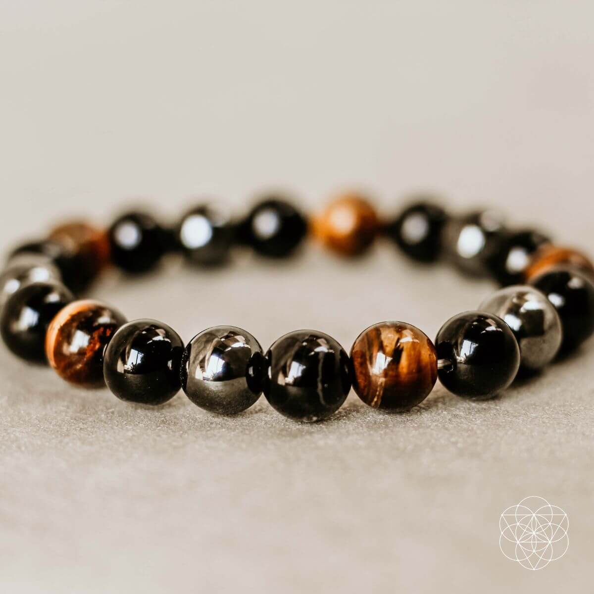 The Triple Protection Bracelet by Conscious Items features glossy black and brown beads, arranged in a circular shape and displayed on a neutral surface. A small white geometric design is visible in the corner.